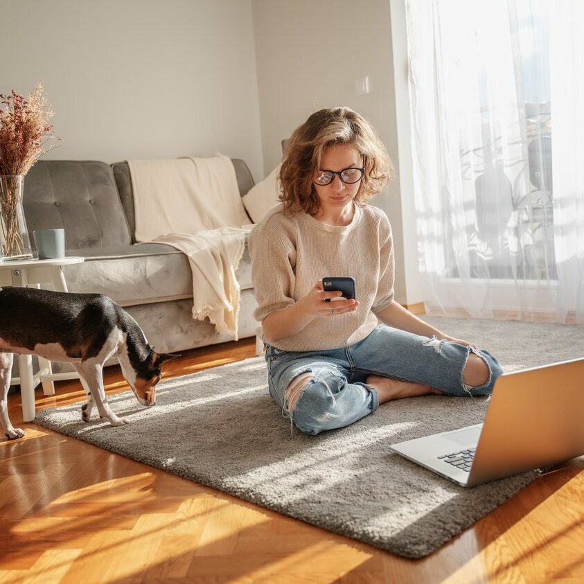 Pretty curly happy young woman sitting at home on the floor in front of laptop with her pet dog working and learning online